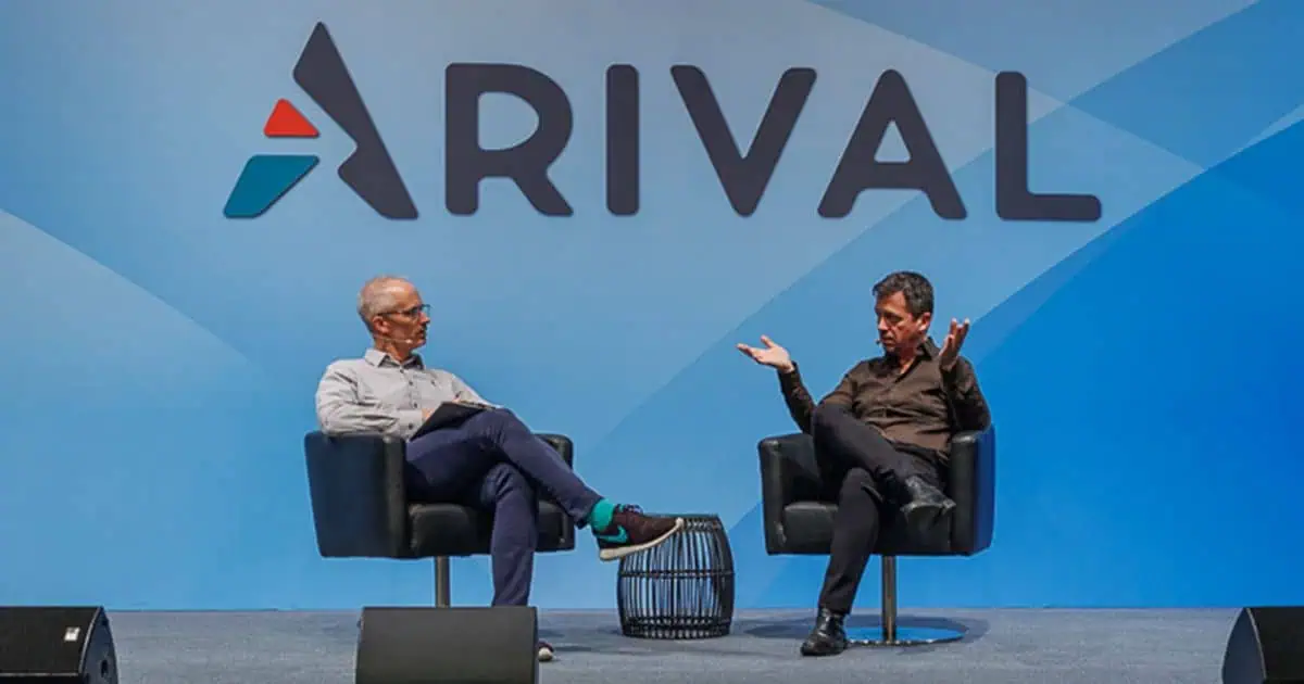 Two people are seated in armchairs on a stage with a large blue background displaying the logo "ARIVAL." The person on the left casually sits with one leg crossed over the other wearing a grey shirt and dark trousers, while the person on the right is gesturing with their hands, dressed in all black. There are speakers on the stage floor in front of them.