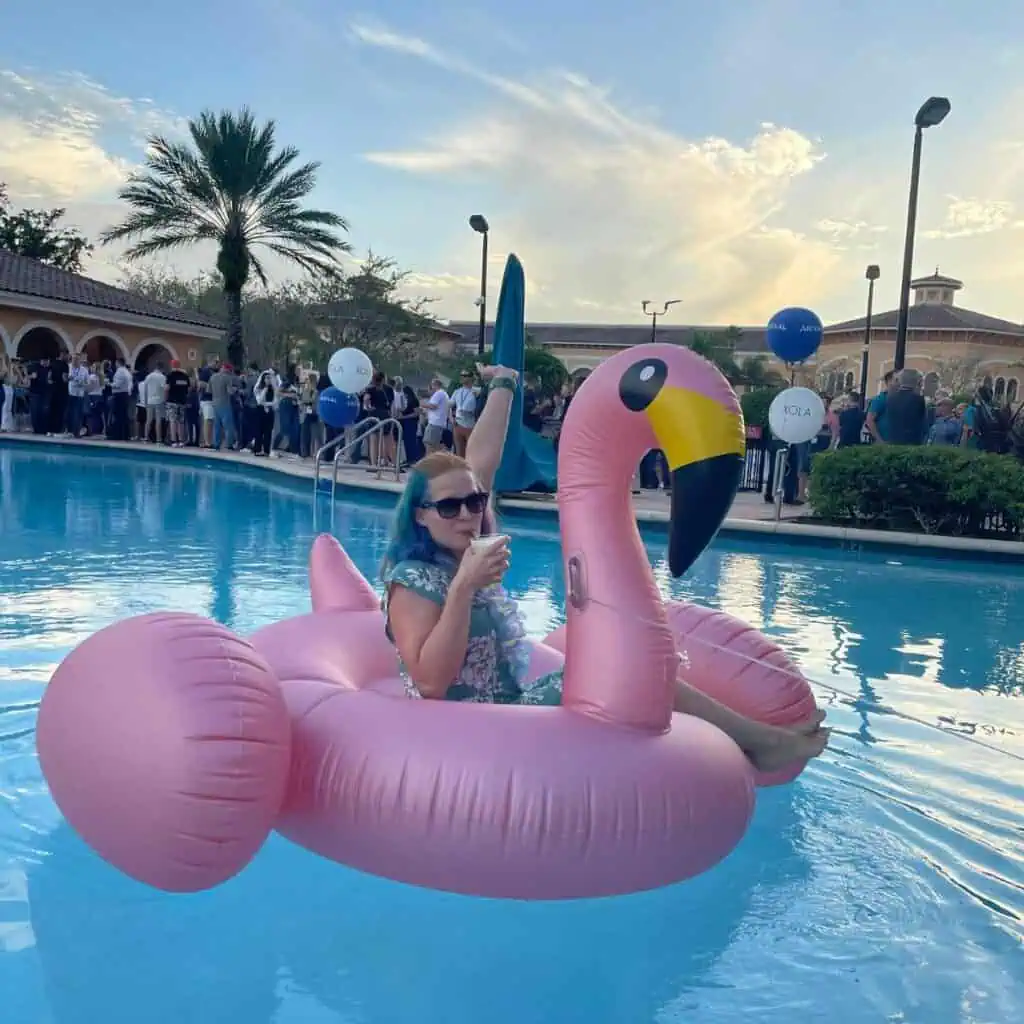 A person lounges on a pink flamingo pool float in a swimming pool, holding a drink, with a blue sky and crowd of people in the background.