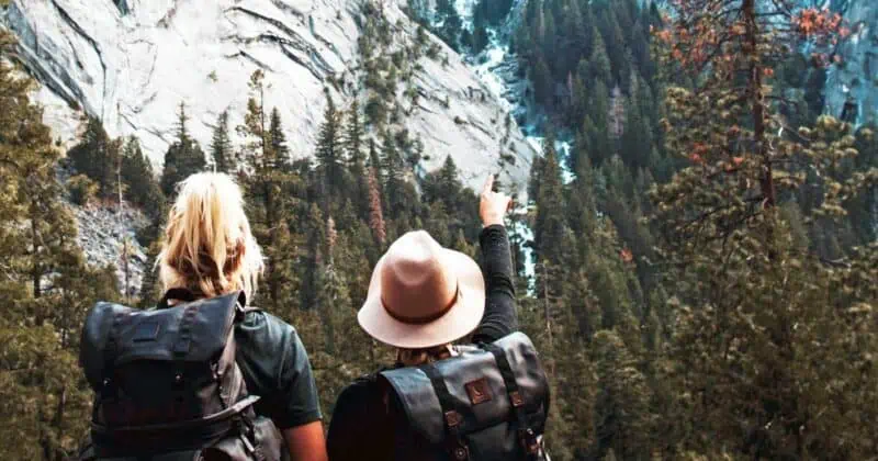 Two hikers with backpacks looking at mountain scenery with trees and rock formations.