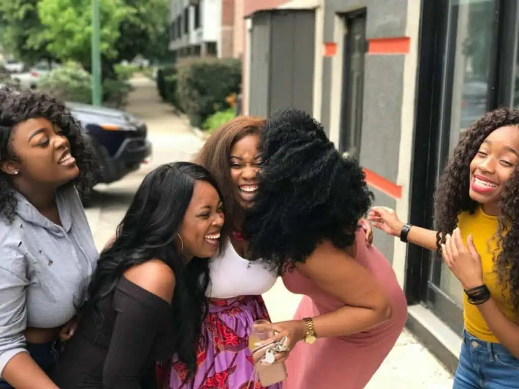 A group of five women laughing and hugging each other on a city street, displaying genuine joy and connection.