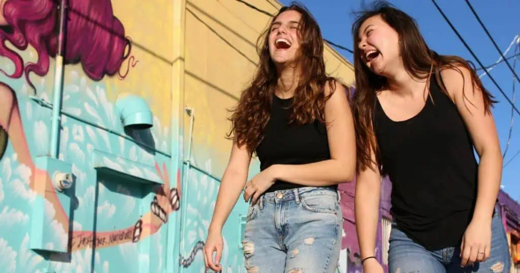 Two young women in black tops and jeans laughing together in front of a colorful mural on a sunny day.