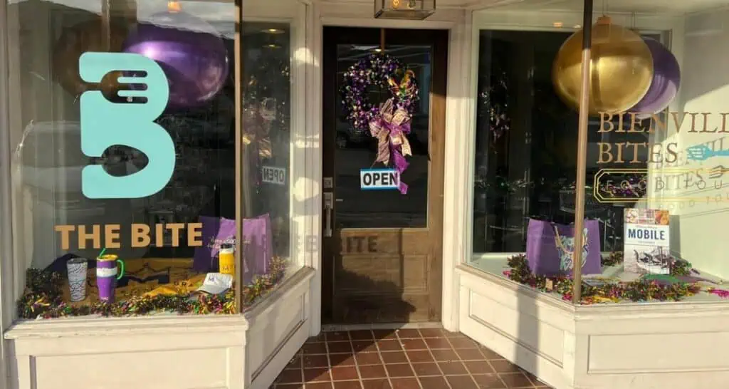 Storefront of "The Bite" restaurant with purple and gold balloons, a festive wreath, and Mardi Gras-themed decorations in the windows.