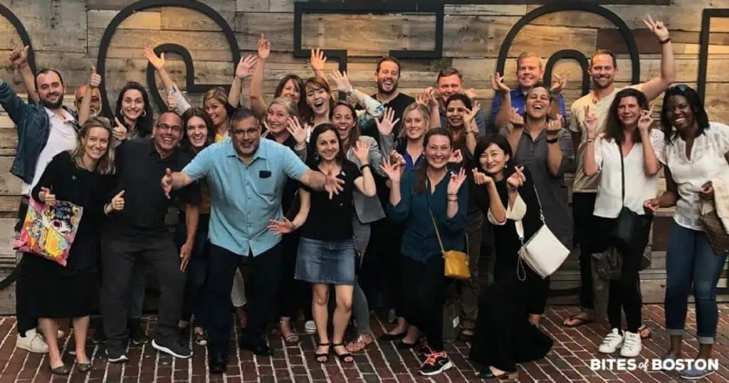 Large group photo of smiling people posing in front of a wooden wall with decorative shapes, labeled "Bites of Boston" at the bottom