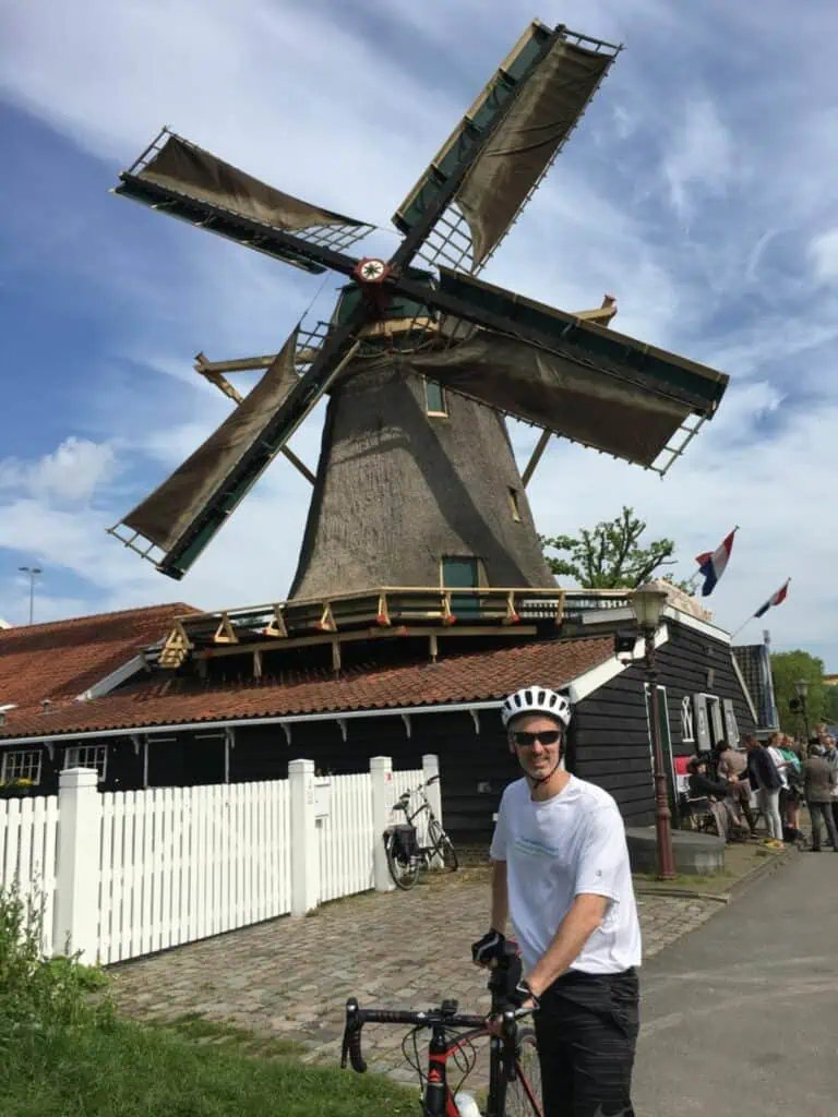 Douglas Quinby wearing a white t-shirt and helmet stands next to a traditional Dutch windmill.