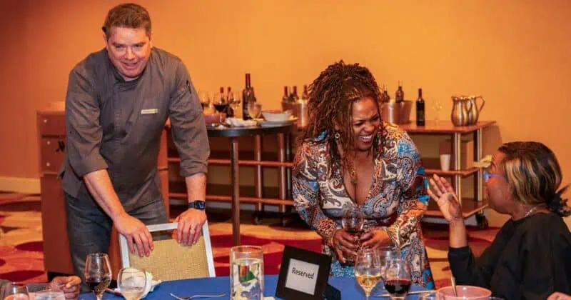 A chef in gray uniform stands at a table with two women, surrounded by wine bottles and glasses in a warmly lit room.