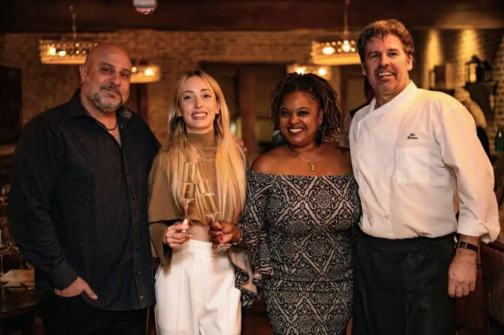Four people, including a chef in white uniform, pose together holding champagne glasses in a dimly lit restaurant setting.