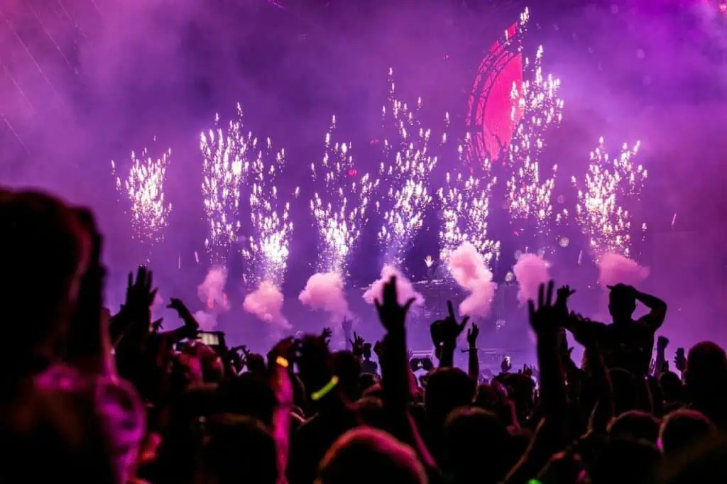 Silhouette of crowd with raised hands at concert with purple lighting and fireworks
