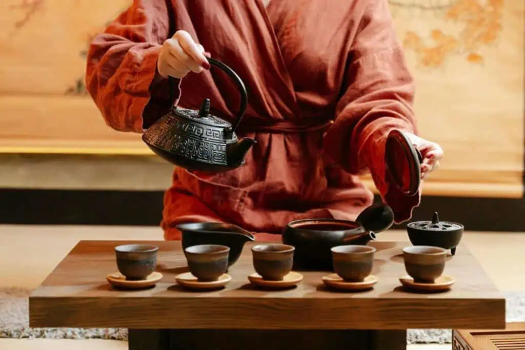Person in red robe pouring tea from ornate teapot into small cups on wooden tray